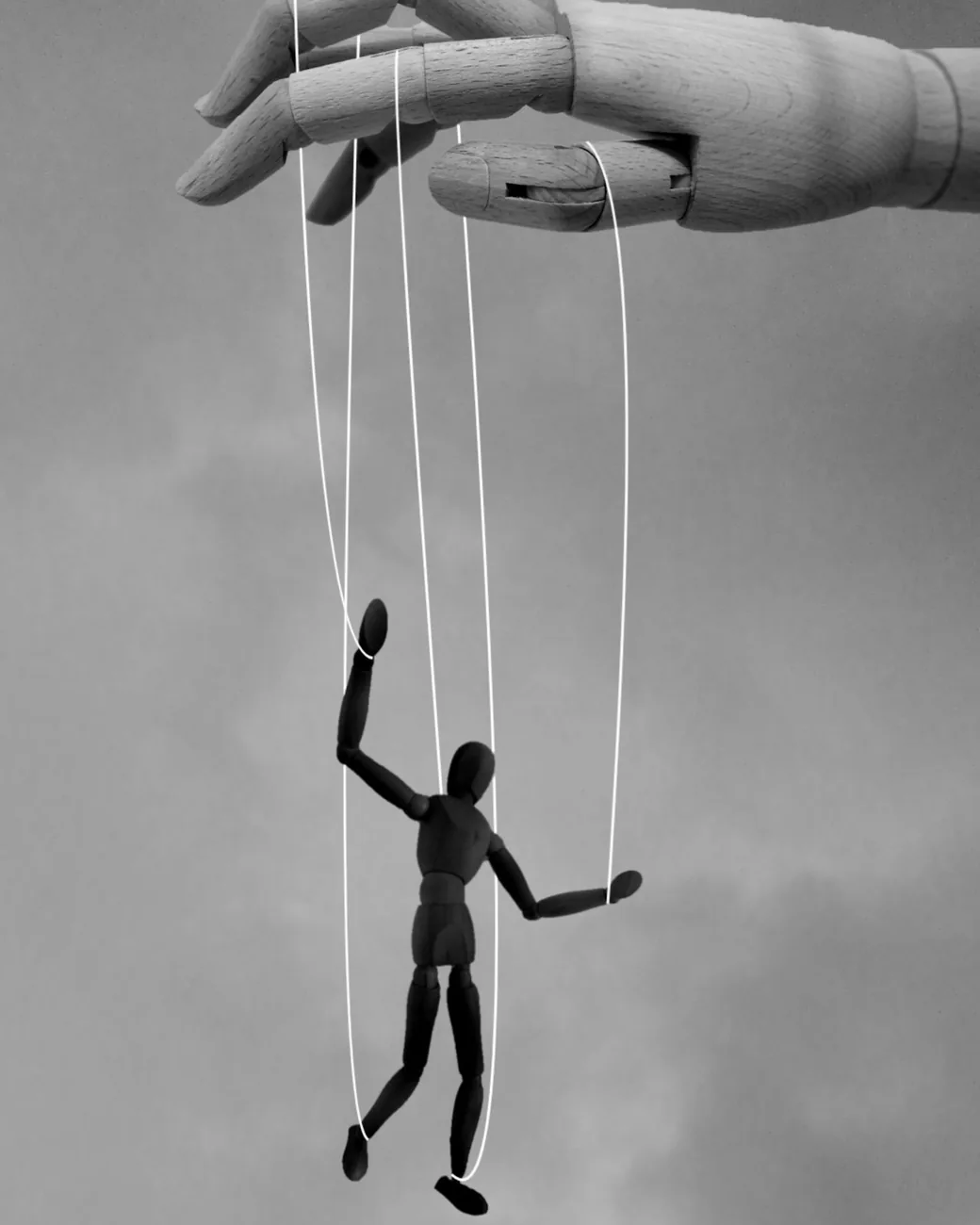 Figura de madera articulada suspendida de hilos y sostenida por una mano de madera sobre un fondo gris neutro.